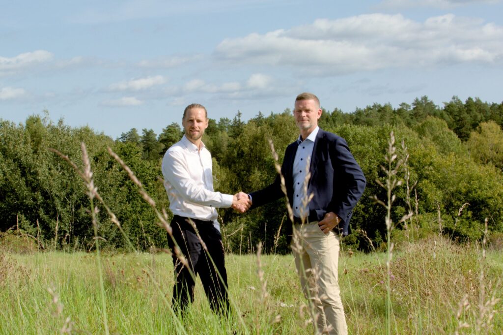 Två män skakar hand på en äng med skog i bakgrunden.