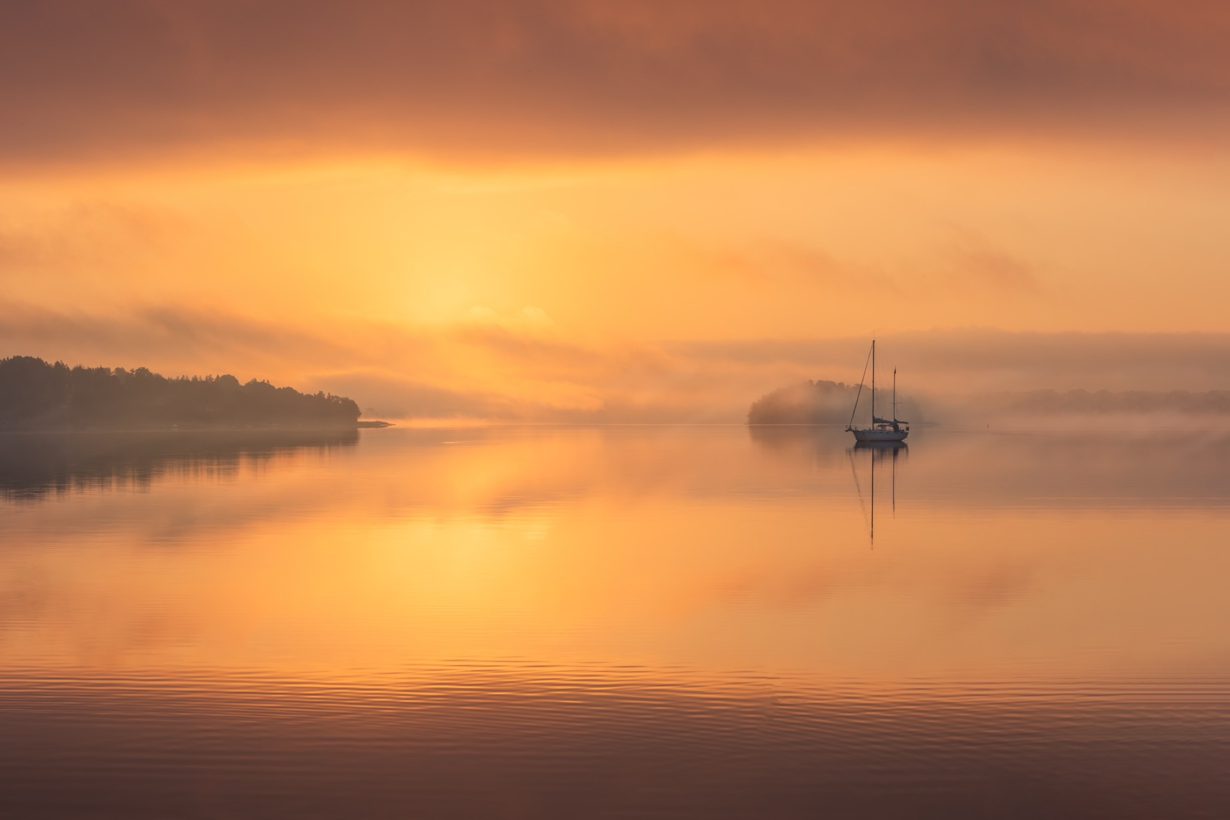 Segelbåt ligger stilla på en spegelblank sjö i soluppgångens varma sken, med dimma som sveper över vattenytan och hav i bakgrunden.