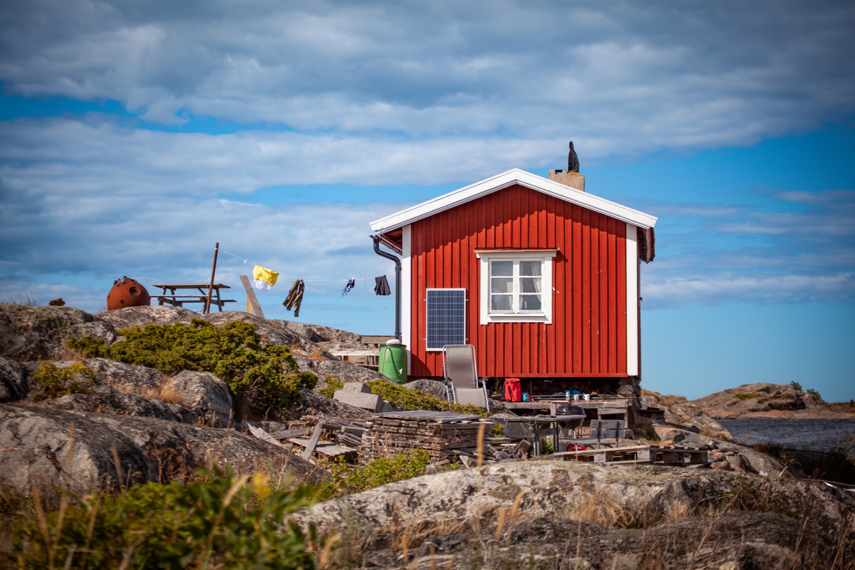 En liten röd stuga med vita knutar står på klippor vid havet under en blå himmel med moln. En solpanel är monterad på väggen, tvätt hänger på lina och omgivningen består av buskar, utemöbler och stenig terräng.