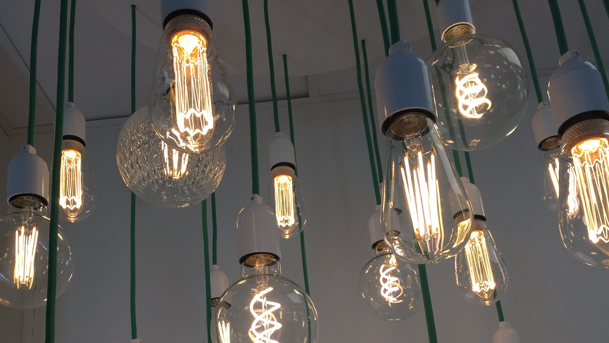 Flera tända glödlampor med olika former och mönster hänger från taket i gröna sladdar. Lamporna lyser med ett varmt sken mot en vit bakgrund.