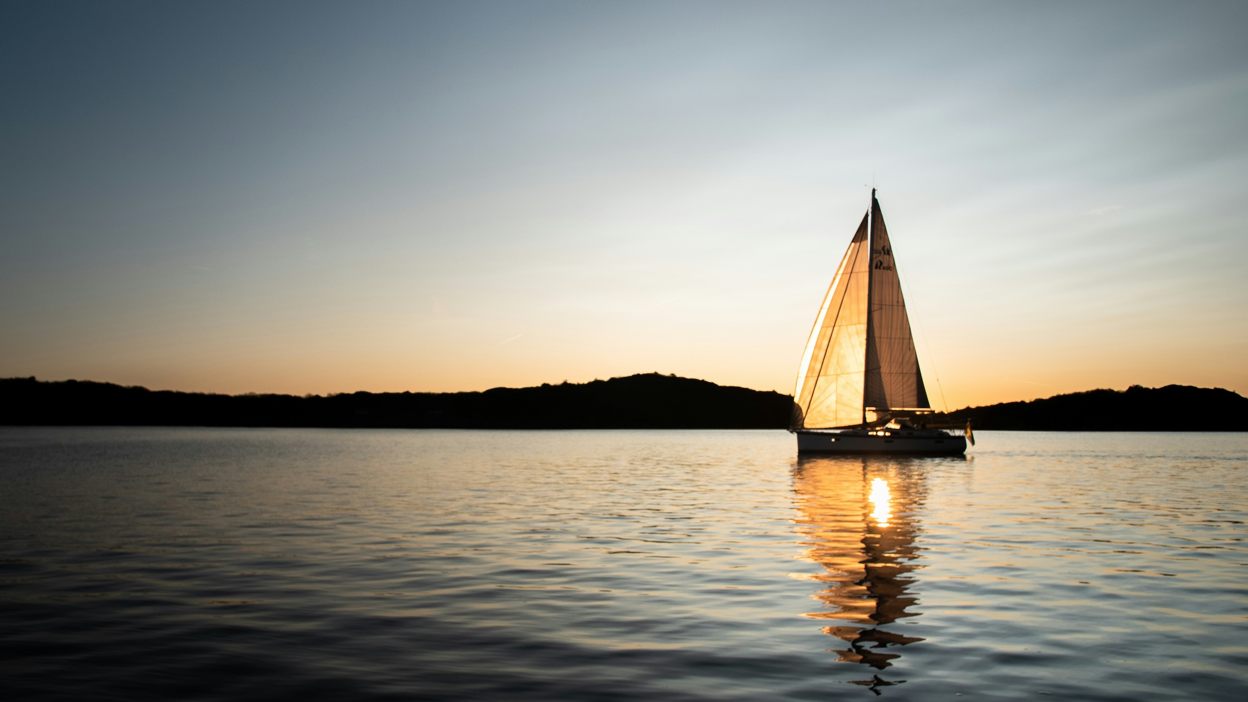 Segelbåt på stilla vatten vid solnedgång, med solens reflex i vattnet och siluett av land i bakgrunden.