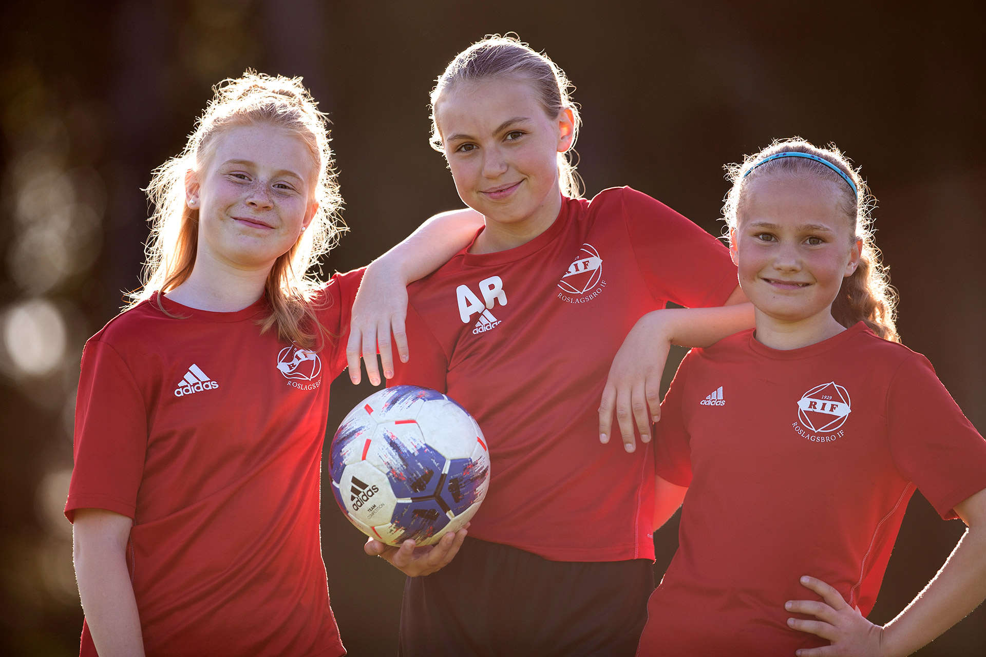 Tre flickor i röda tröjor står tillsammans. En av dem håller i en fotboll.