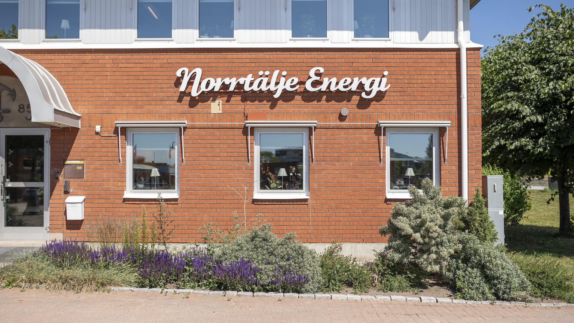 Fasaden på Norrtälje Energis kontor i rött tegel med vit logotyptext ovanför tre fönster, omgiven av planteringar med blommor och buskar.