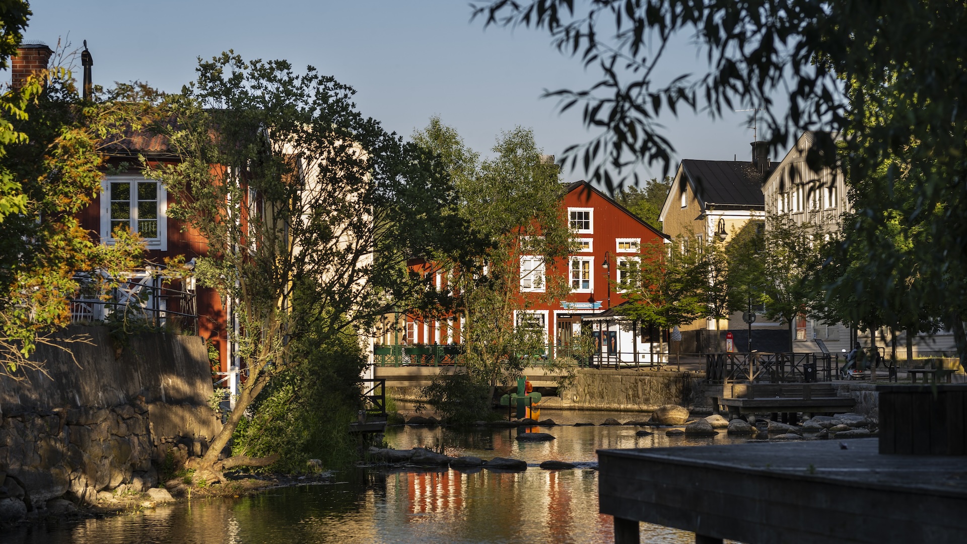 Vy över åpromenaden i kvällssol med röda och vita trähus längs vattnet i Norrtälje. Träd ramas in i förgrunden och vattnet speglar husfasaderna.