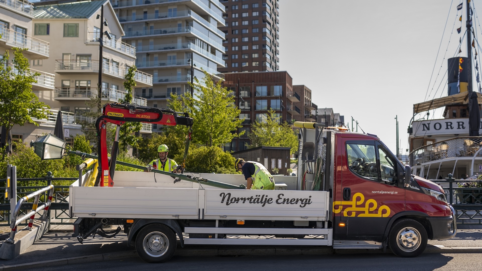En solbelyst småstadsgata (Norrtälje) kantad av trähus, butiker och apotek, med skogsklädd höjd i bakgrunden. Två arbetande män från Norrtälje Energi och en företagsbil med kran.