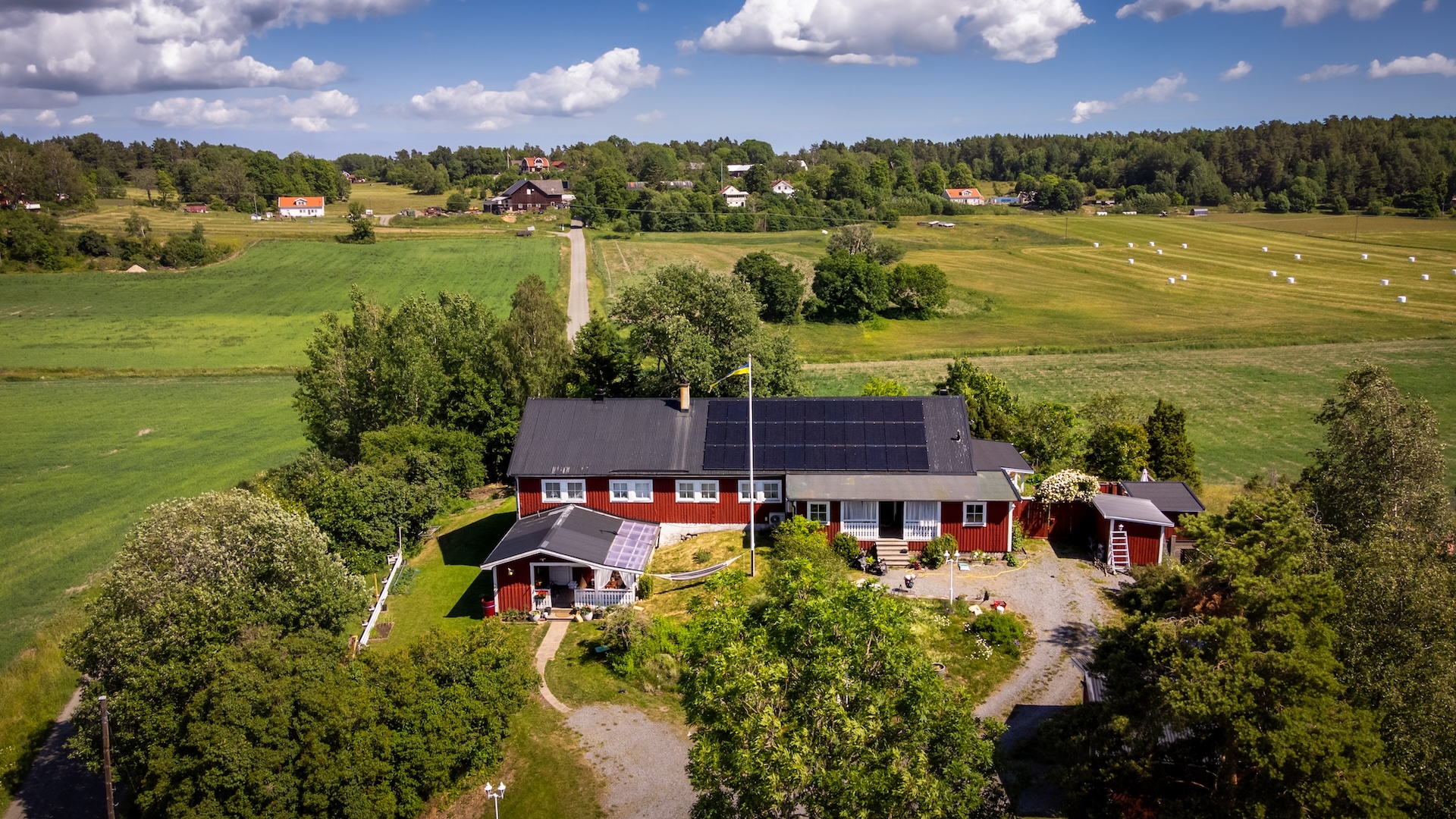 Bild på hus med solceller på taket. Flygfoto över ett rött hus med svarta solceller på taket, omgivet av grönska, grusväg och öppna fält under en blå himmel.