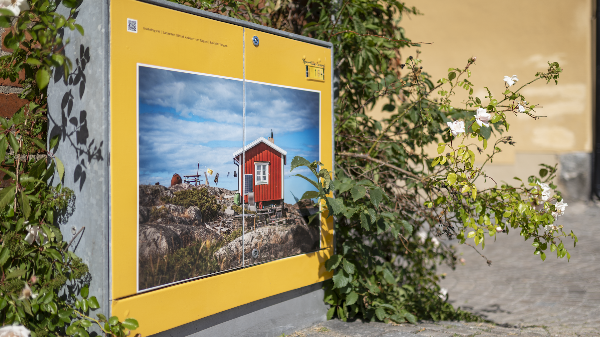 Ett elskåp med konstmotiv av ett rött skärgårdshus, inramat av gul kant och omringat av rosor.