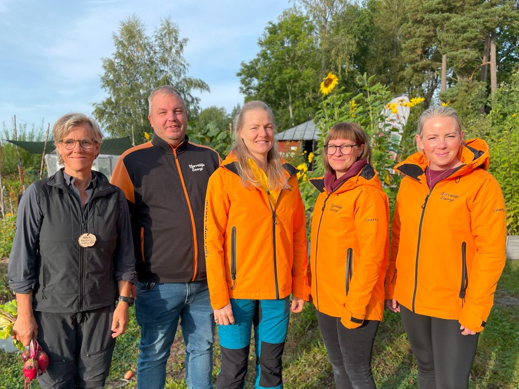 Fem personer står tillsammans utomhus framför solrosor och grönska. Fyra av dem bär orangea jackor med Norrtälje Energi-logga, en person till vänster bär mörka kläder och håller i grönsaker.