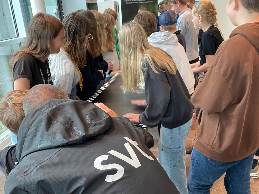 Grupp barn och ungdomar samlade runt aktivitet vid ett bord, en person i SVT-jacka i förgrunden.