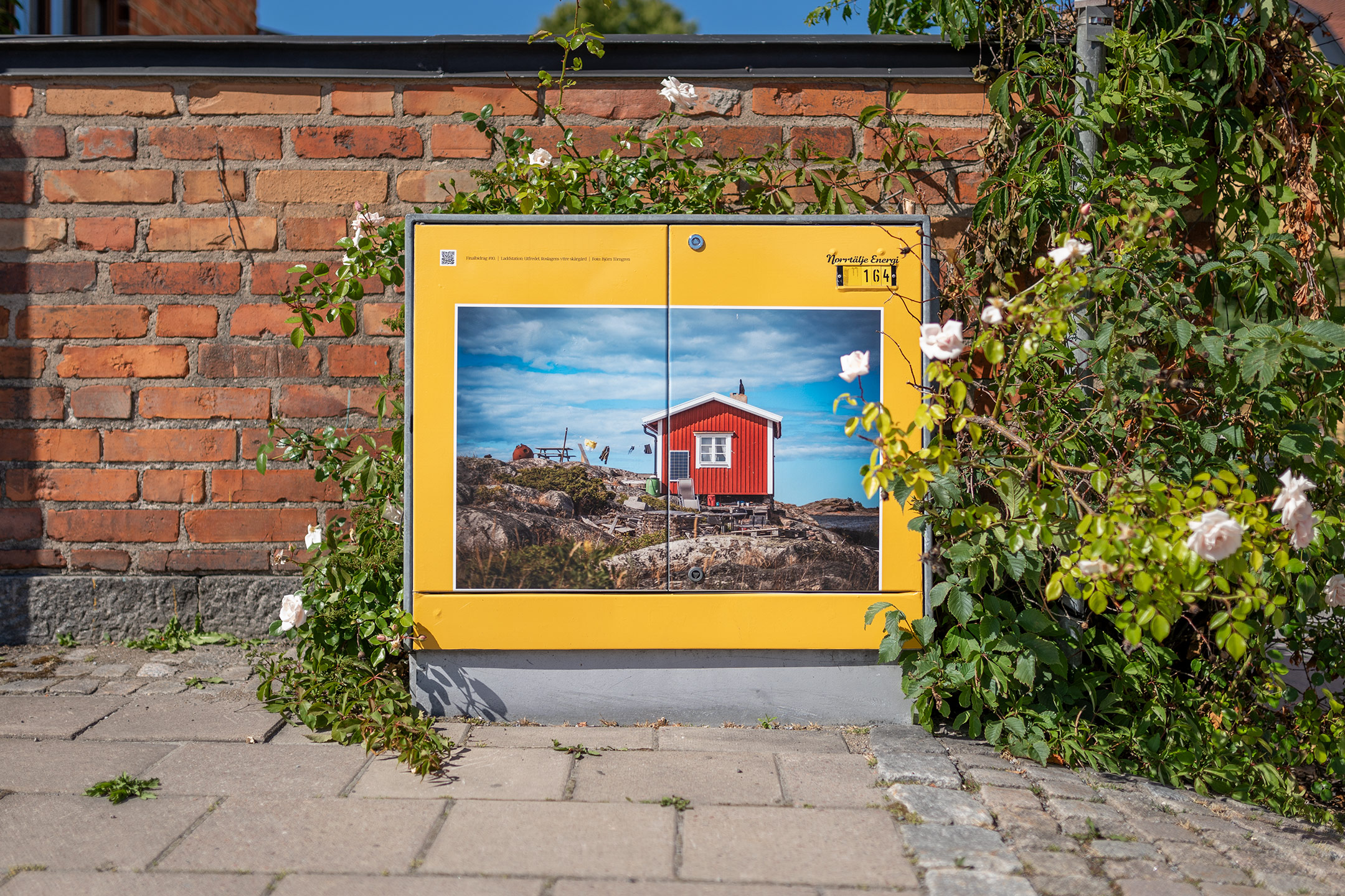 Ett elskåp med konstmotiv av ett rött skärgårdshus, inramat av gul kant och omringat av rosor.