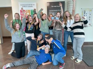 En stor grupp barn och ungdomar poserar glatt i en ljus inomhusmiljö. Många av dem har sina händer i luften och visar tecken på glädje och firande. De bär avslappnade kläder och har medaljer runt halsen, vilket antyder att de deltagit i en tävling eller ett evenemang. På väggen bakom gruppen finns affischer med texten 'Gemensam kraft för ett hållbart samhälle' och 'Norrtälje Energi'
