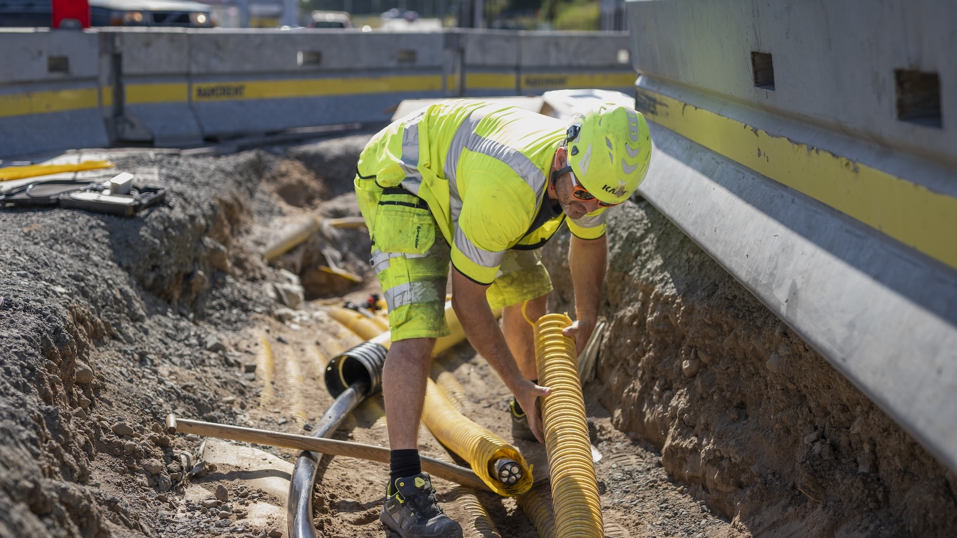 En eltekniker i varselkläder, hjälm och skyddsglasögon arbetar vid en vägarbetsplats. Han hanterar en gul kabelkanal.
