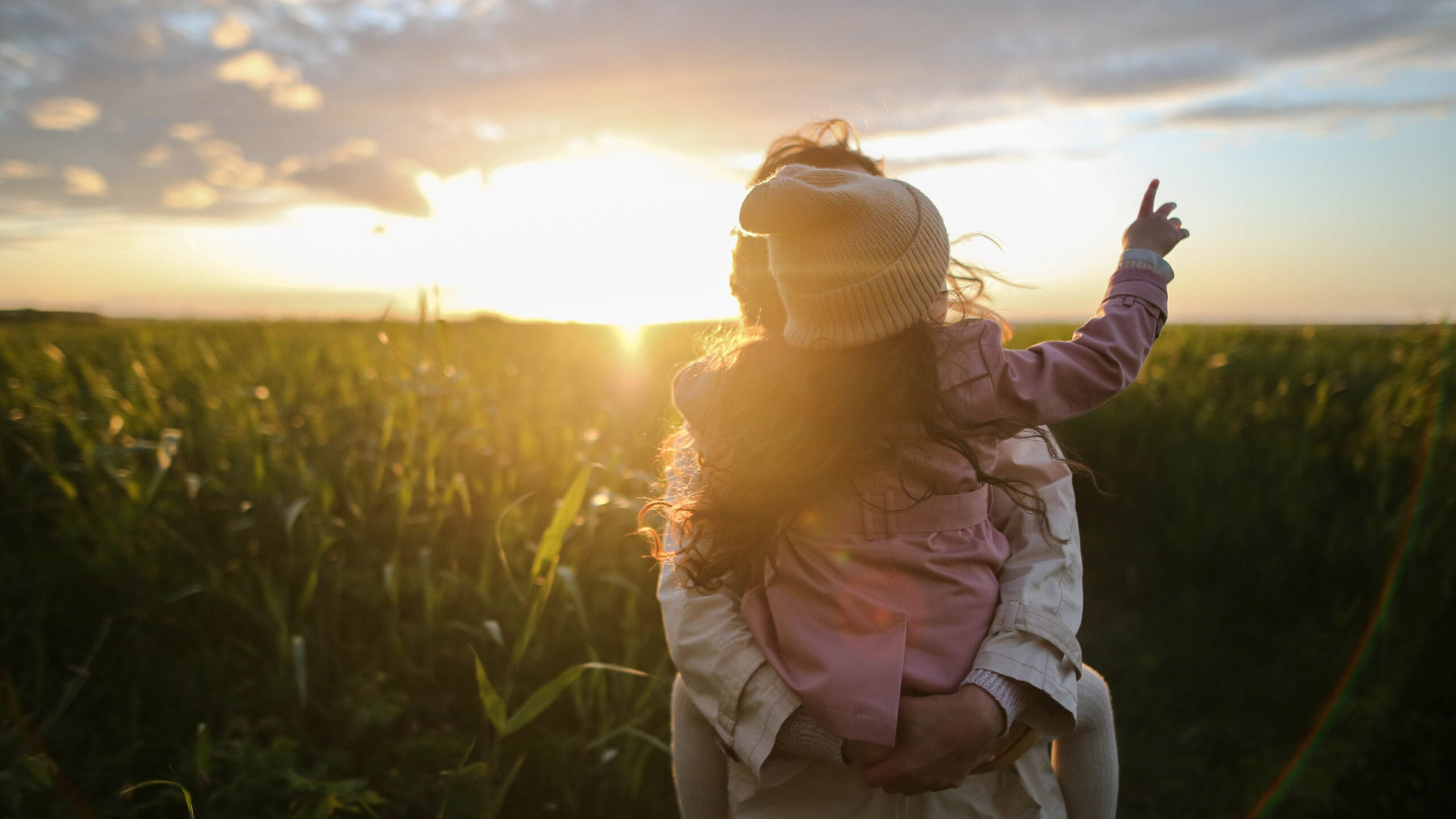 En vuxen som håller ett barn i famnen. De är vända mot varandra i en solnedgång med ett fält av grönt i bakgrunden.