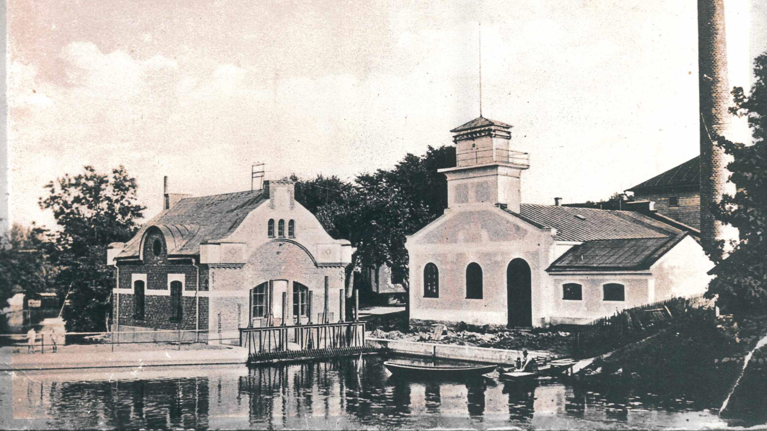 Foto på Norrtälje Elektricitetverk. Historiskt fotografi av två byggnader vid vattnet.