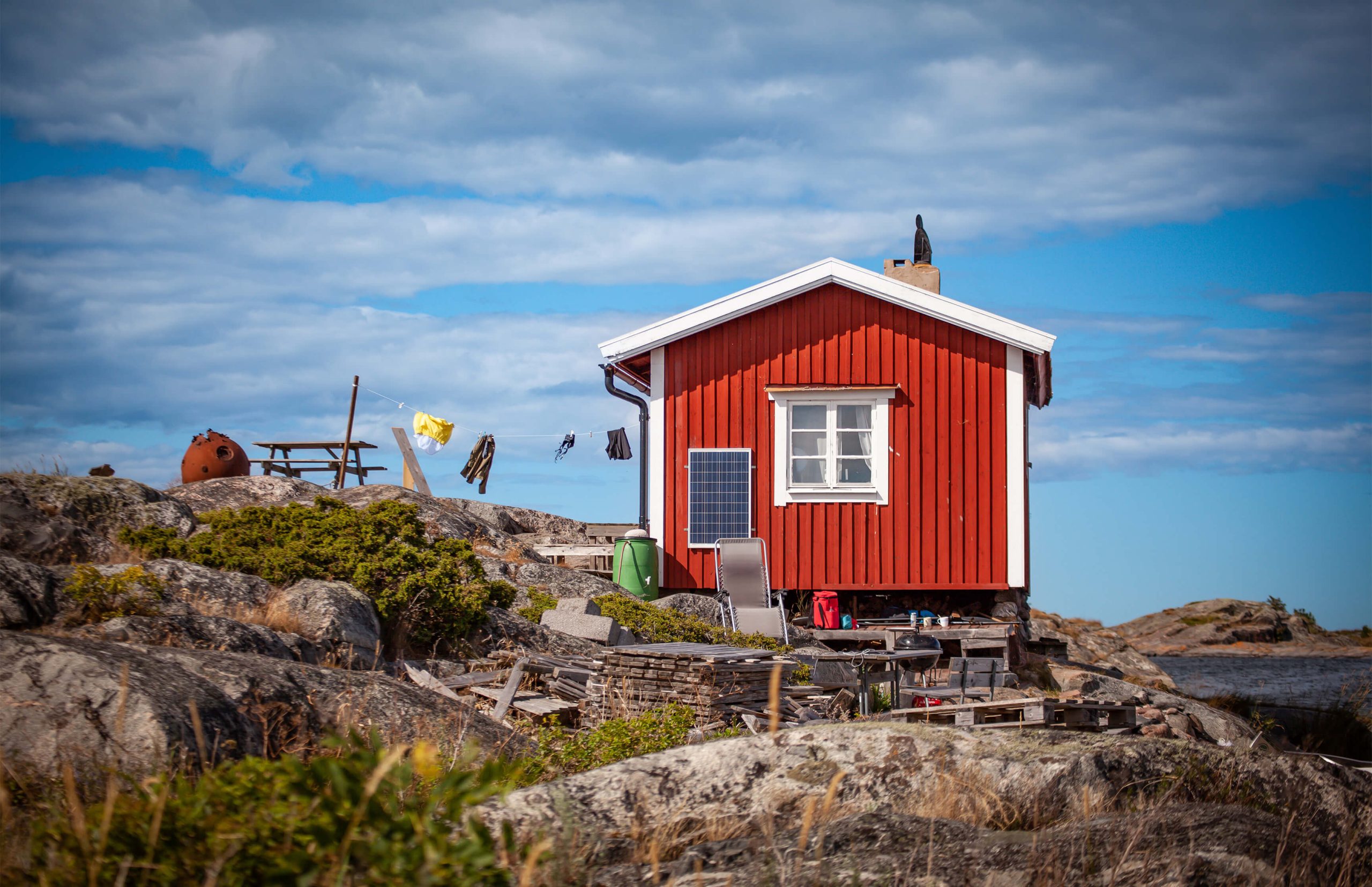 En liten röd stuga med vita knutar står på klippor vid havet under en blå himmel med moln. En solpanel är monterad på väggen.