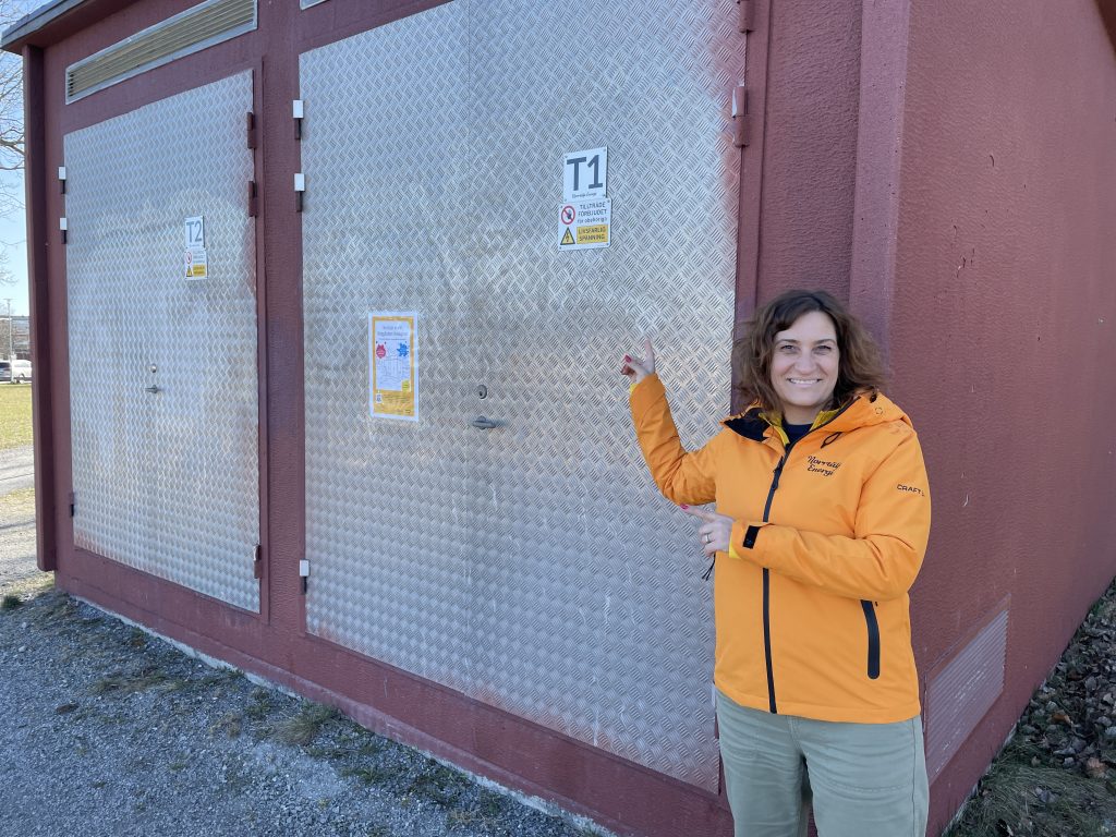 En kvinna i gul jacka med texten "Norrtälje Energi" står leende framför en nätstation med metalliska dörrar och röda väggar.