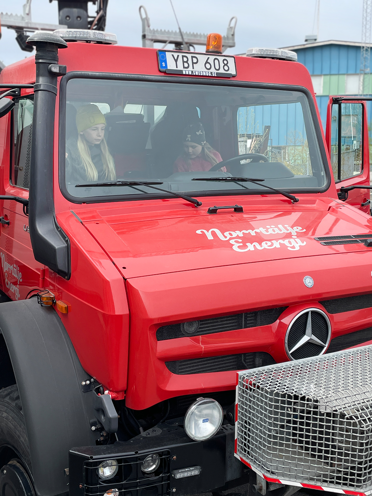 Två barn sitter i förarsätet på en röd arbetsbil märkt “Norrtälje Energi”. De ser koncentrerade ut medan de utforskar fordonet.