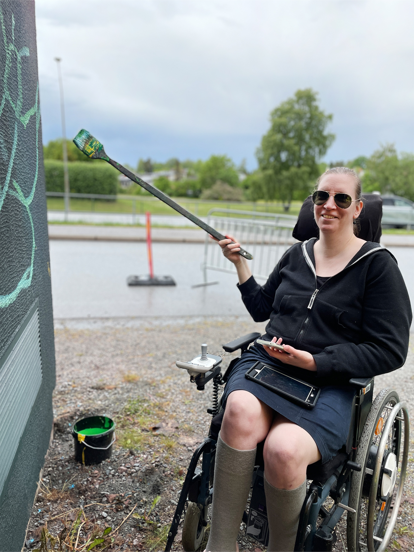 En kvinna i rullstol målar på en vägg med en lång pensel. Hon bär solglasögon och en svart hoodie, och har en mobiltelefon i ena handen. En hink med grön färg står på marken bredvid henne. I bakgrunden syns en väg, grönska och ett träd under en mulen himmel.