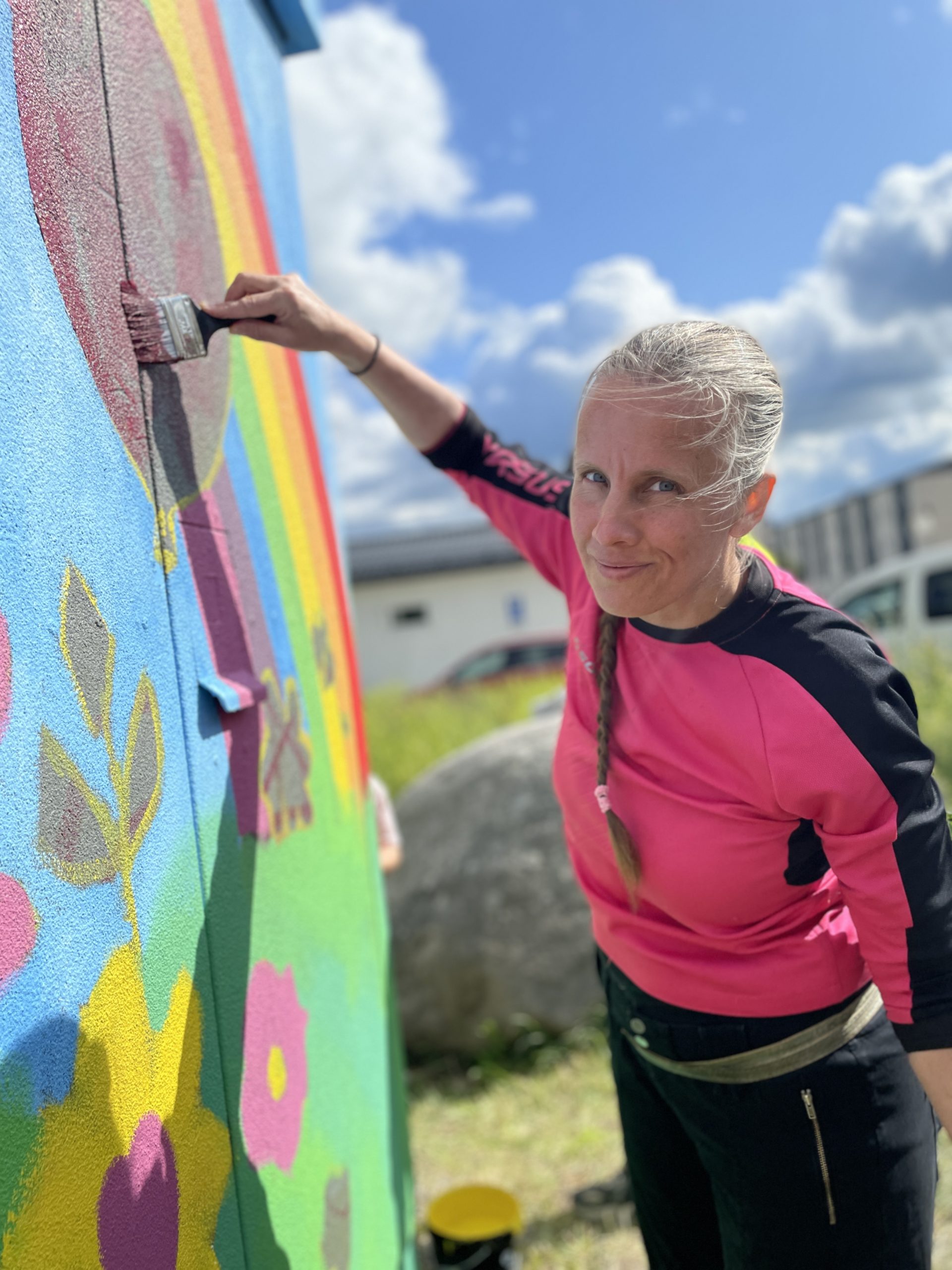 En kvinna i rosa tröja målar en färgglad mural med regnbågsmotiv och blommor på en vägg utomhus. Hon håller en pensel i handen och ser mot kameran med ett leende. Det är soligt väder med blå himmel och moln, och i bakgrunden syns gräs, en sten och en byggnad.