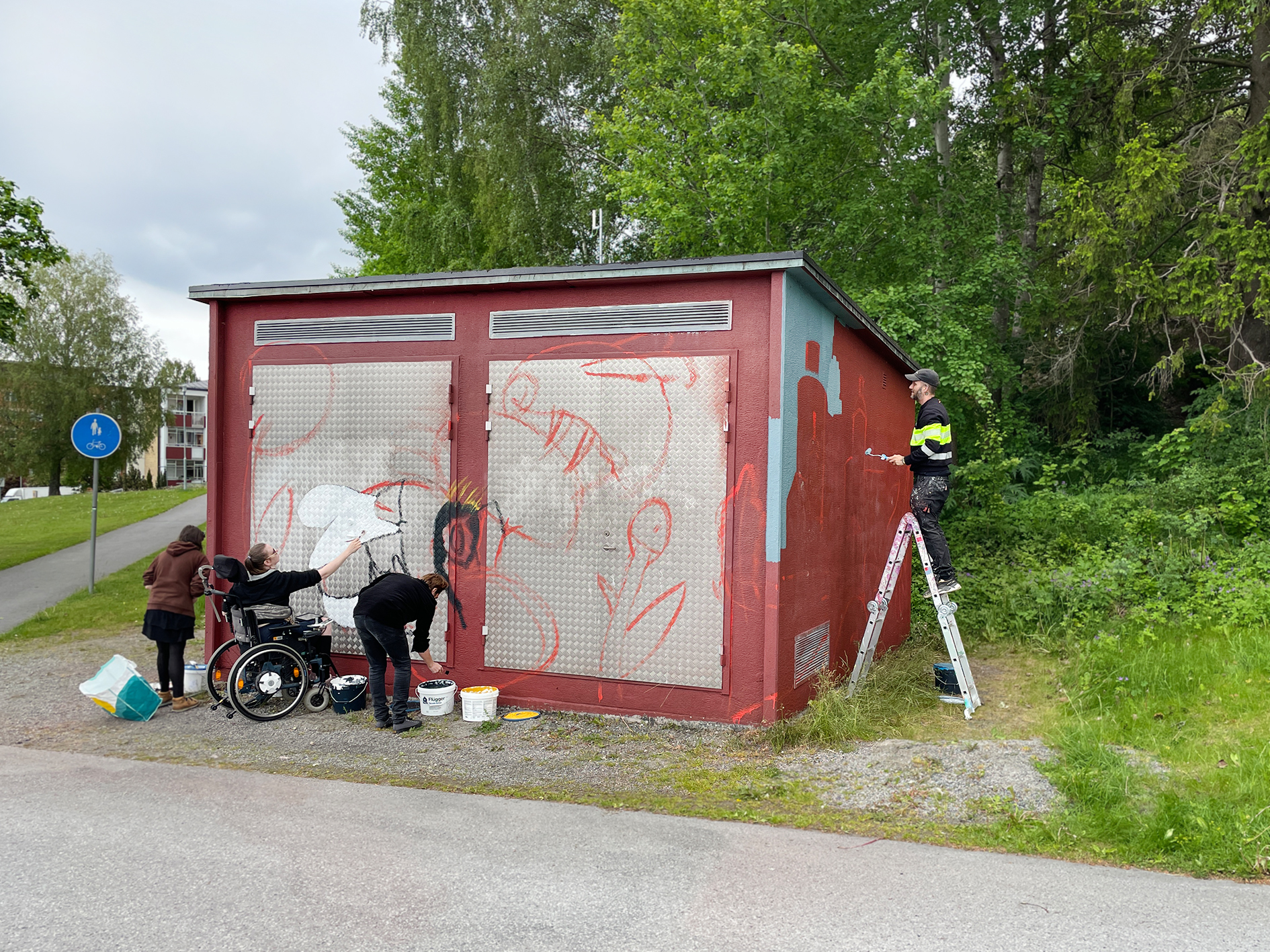 Fyra personer målar en röd byggnad utomhus. En person i rullstol målar tillsammans med två andra personer på byggnadens framsida, där konturerna av en muralmålning är skissade.