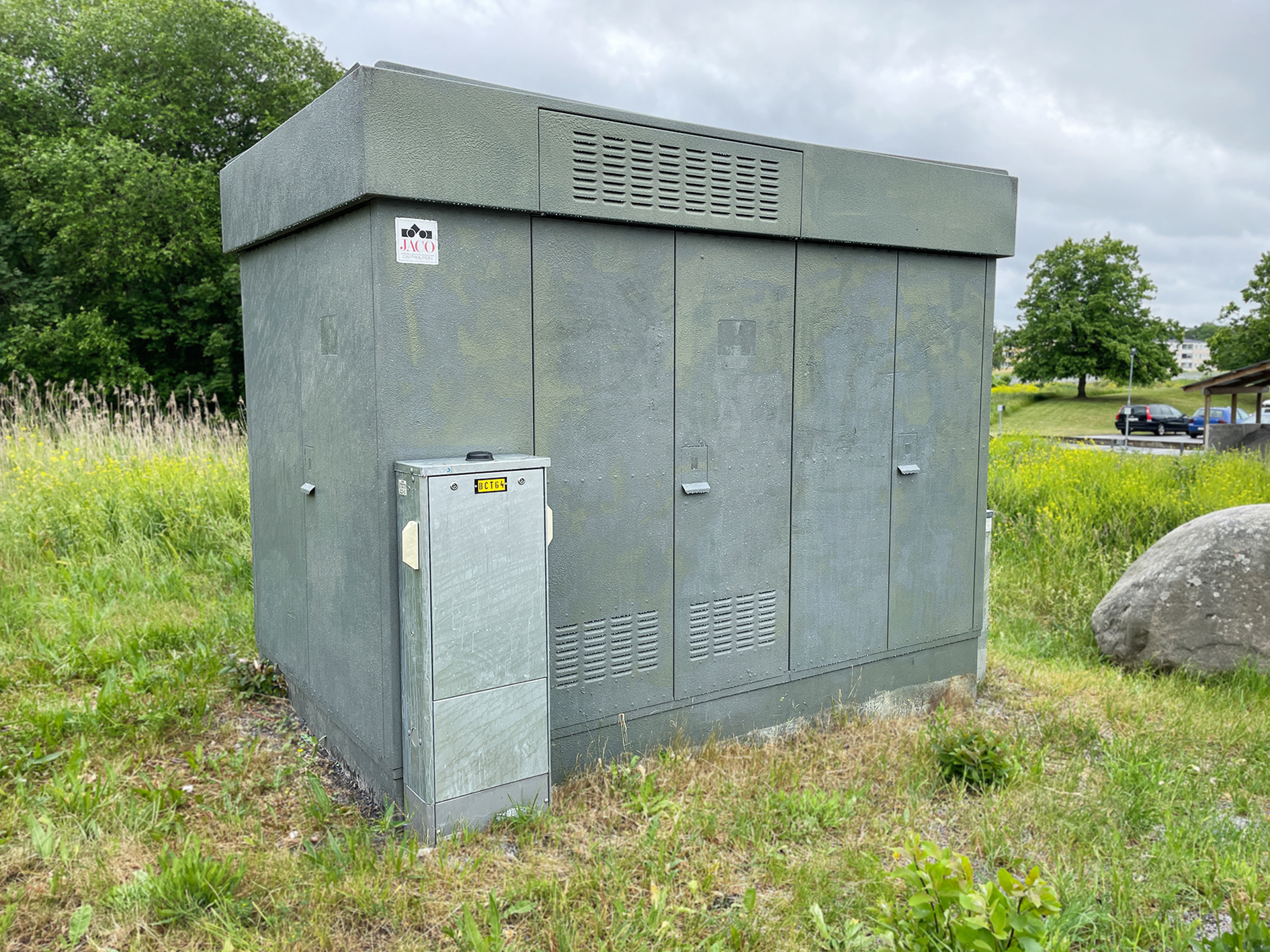En grön nätstation i metall står på en grästäckt yta. Bakom den syns högt gräs, träd och en parkering med några bilar.