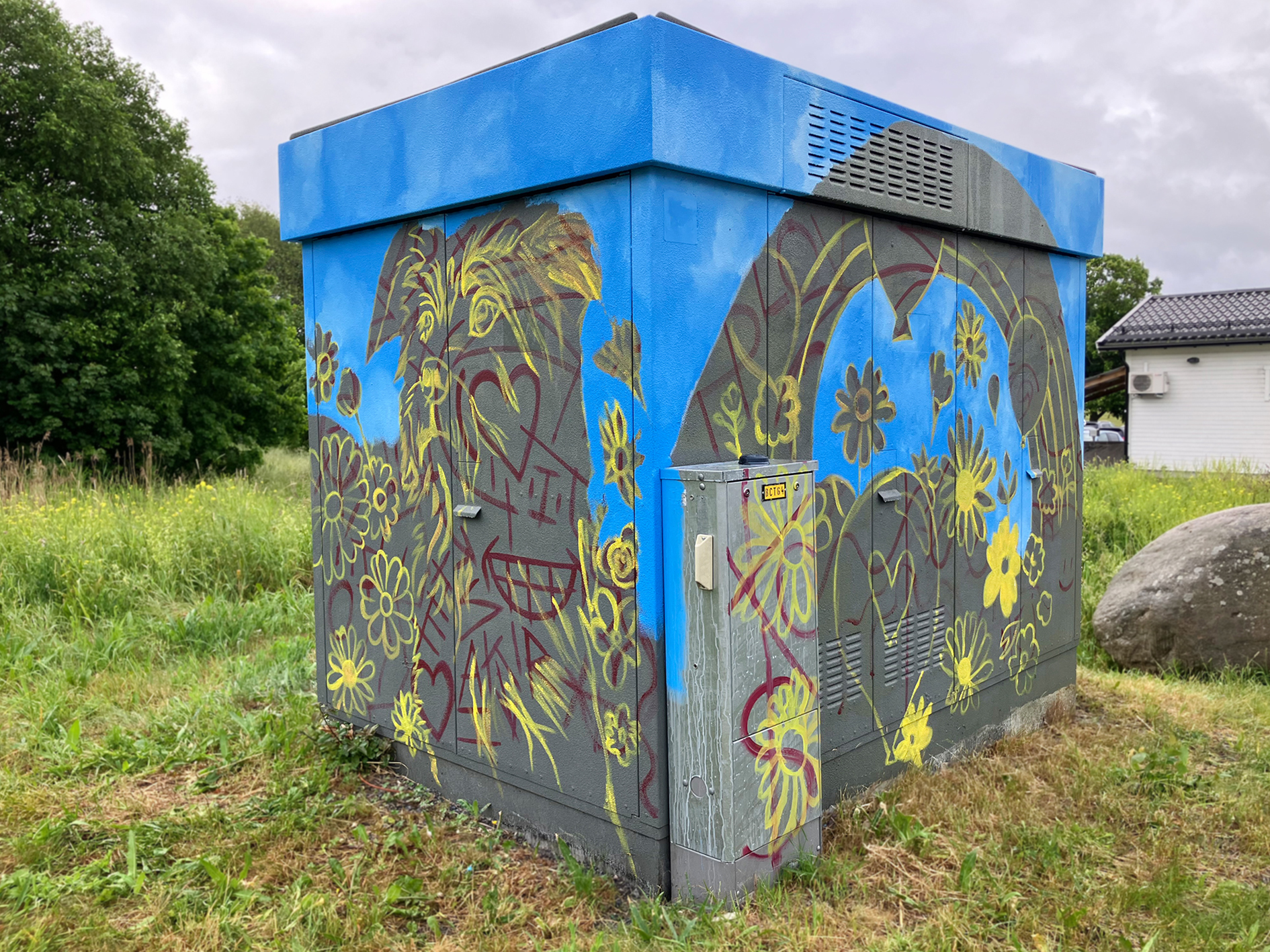 En elstation på Vigelsjövägen som har börjat målas med ett färgstarkt motiv. Bakgrunden är delvis blå, och över ytan syns skisser av gula blommor och en glad ansiktsfigur i rött.