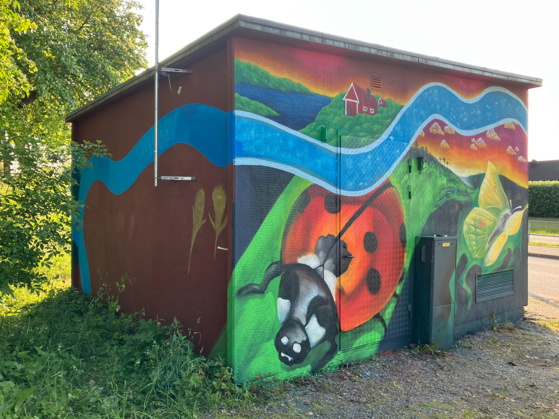 En nätstation, är täckt av en färgstark muralmålning. På ena sidan syns två stora humlor som pollinerar lila och orange blommor, omgivna av grönska och fler blommor.