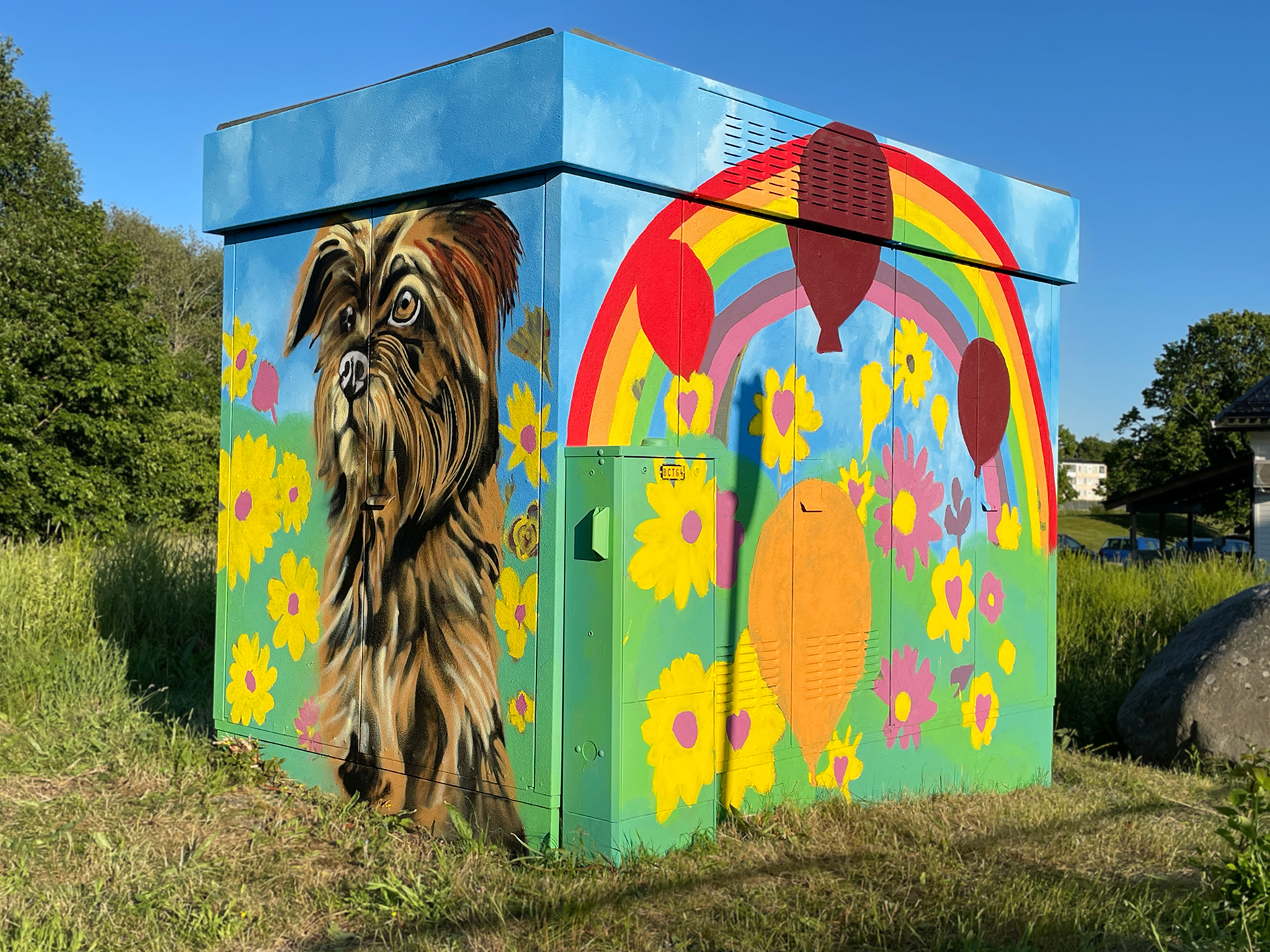 Ett elskåp utomhus målat i starka färger. På framsidan syns ett realistiskt porträtt av en liten långhårig hund, omgiven av gula blommor.