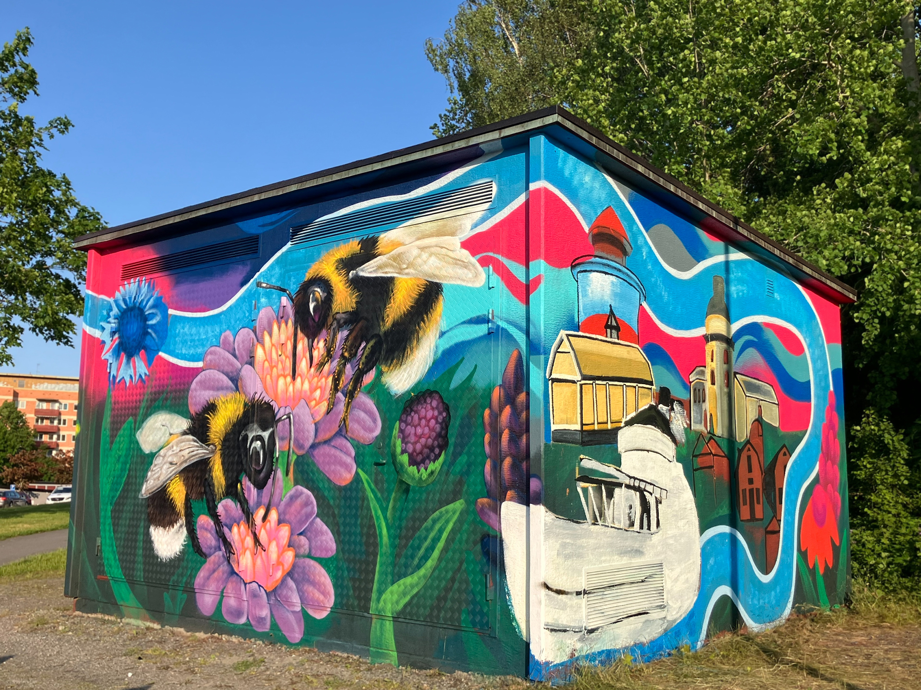 En nätstation, är täckt av en färgstark muralmålning. På ena sidan syns två stora humlor som pollinerar lila och orange blommor, omgivna av grönska och fler blommor.