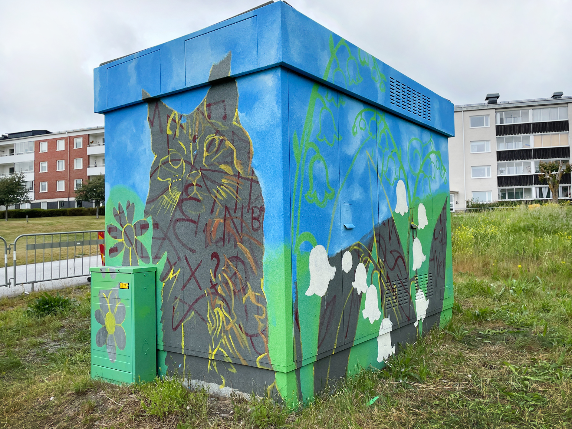 En elnätsstation är dekorerad med färgglad muralmålning som föreställer en grå katt, vita liljekonvaljer och blommor mot en blå himmel.