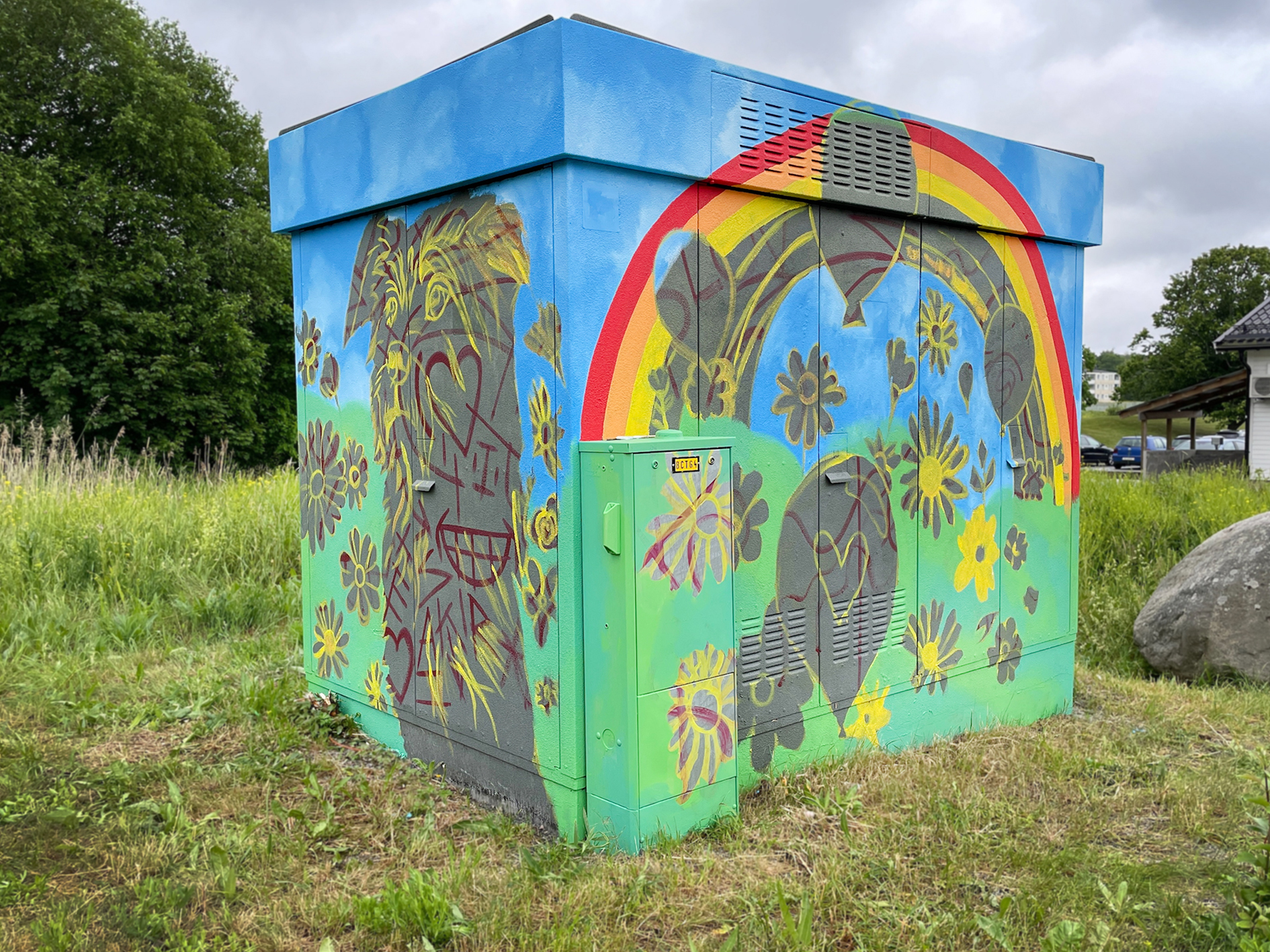 En elnätsstation är målad med färgglad gatukonst som föreställer blommor, hjärtan, ballonger och en regnbåge mot en blå himmel.