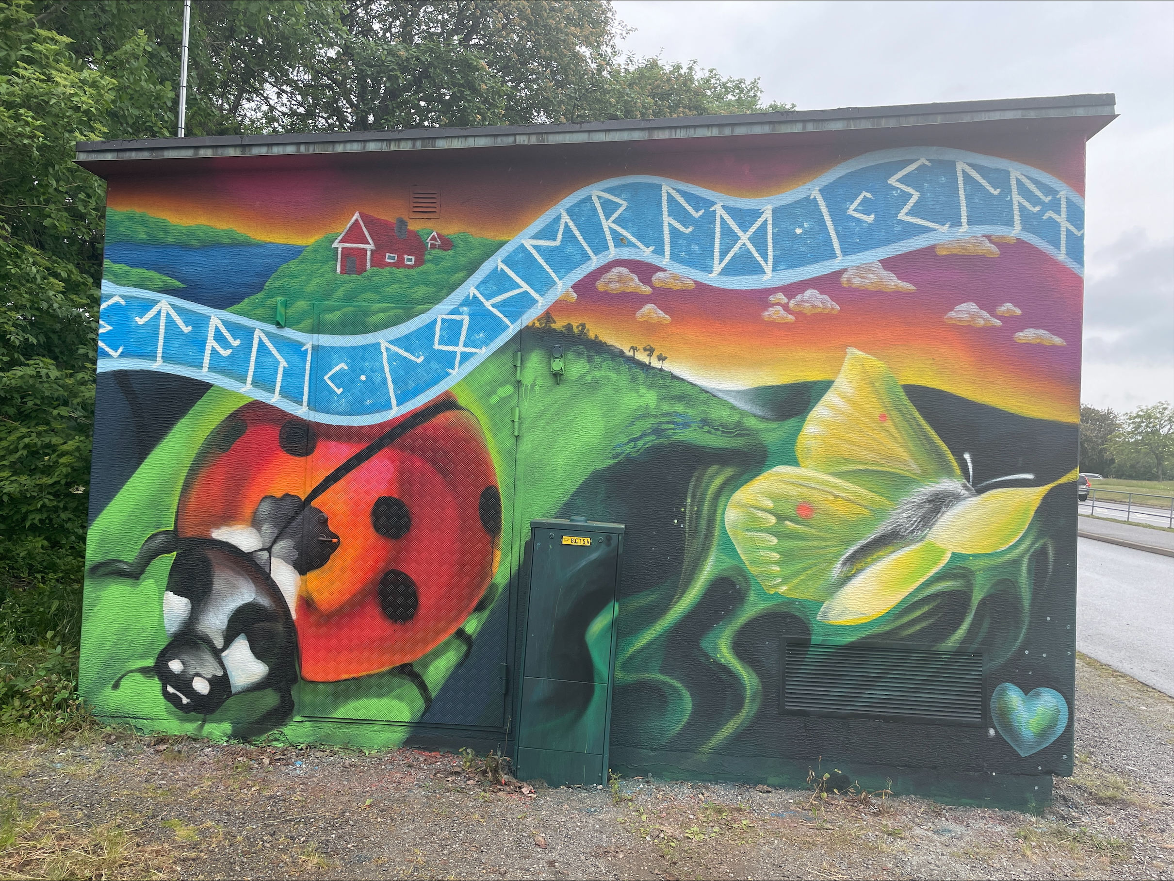 Färgglad muralmålning av en nyckelpiga och en gul fjäril mot en böljande grön landskapshorisont med solnedgång i bakgrunden.