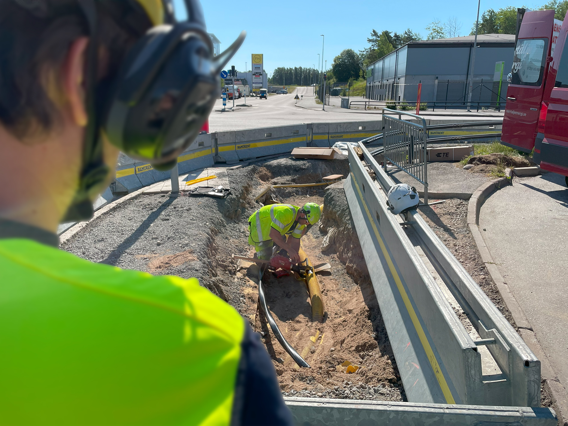 En person med väst som tittar på en eltekniker som arbetar i varselkläder ute.