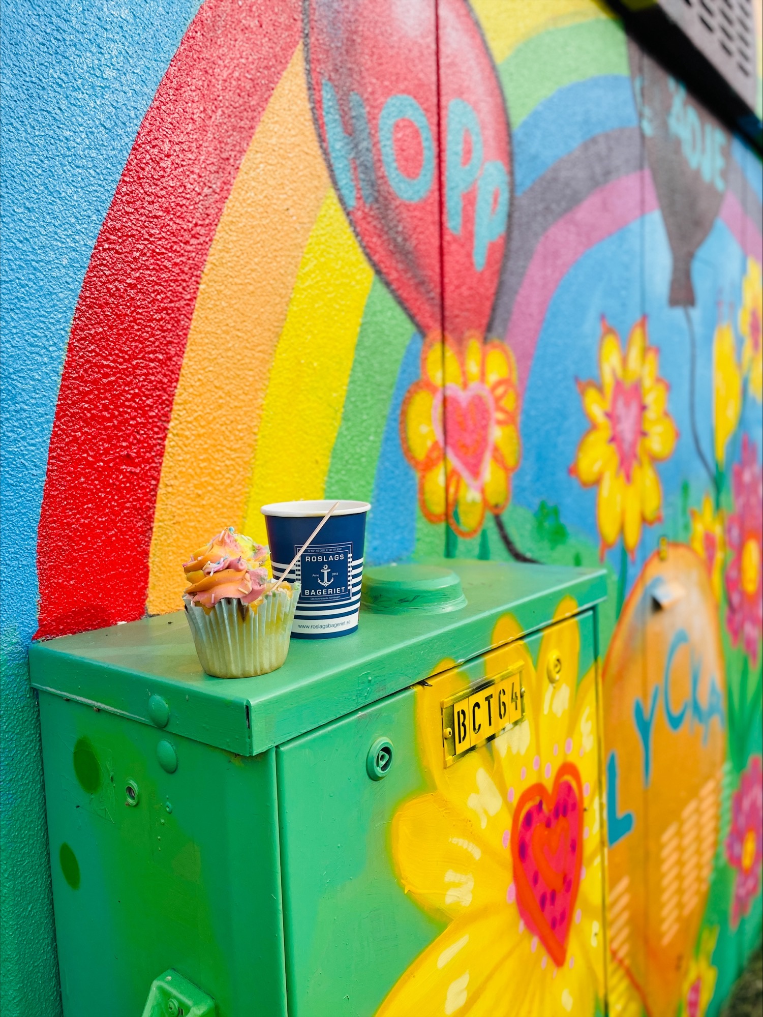 Kopp och cupcake på elskåp framför färgglad muralmålning med regnbåge.