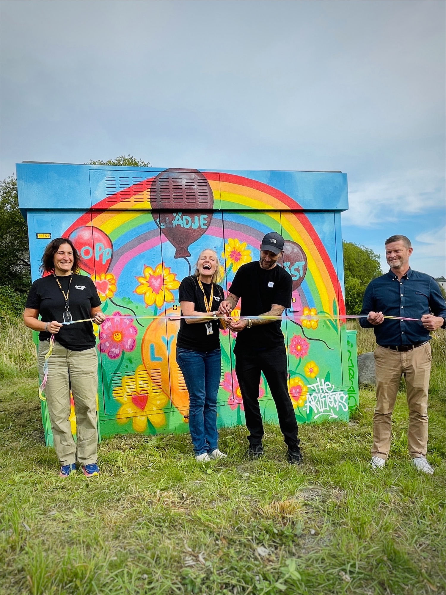 Invigning framför färgglad konstnärlig nätstation med regnbåge och blommor.