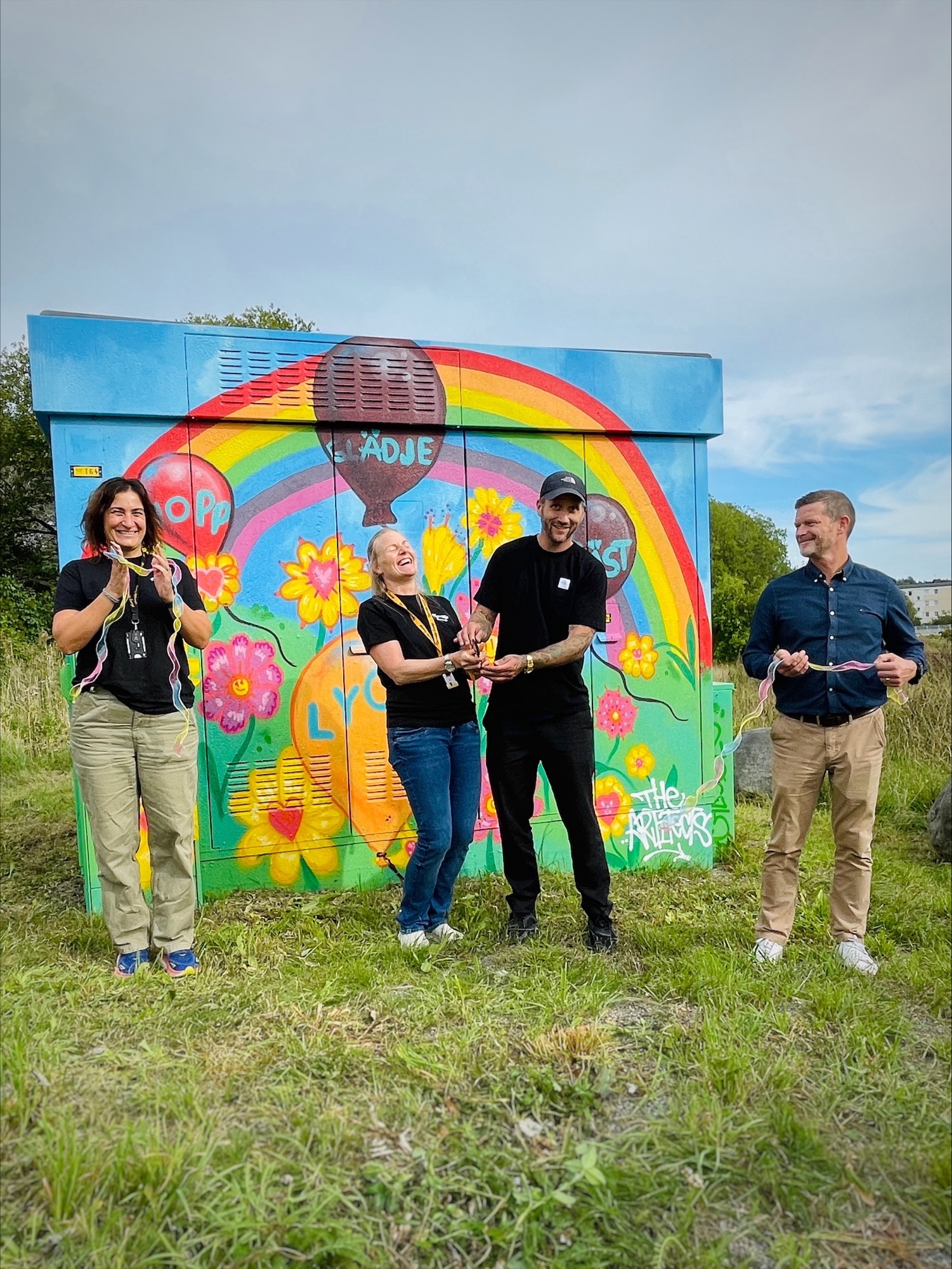 Personer vid invigning framför färgglad konstnärlig nätstation med regnbåge och blommor.
