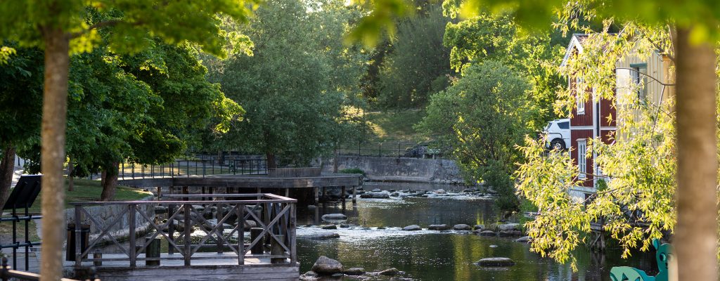 Å med träd, brygga och hus i kvällssol.