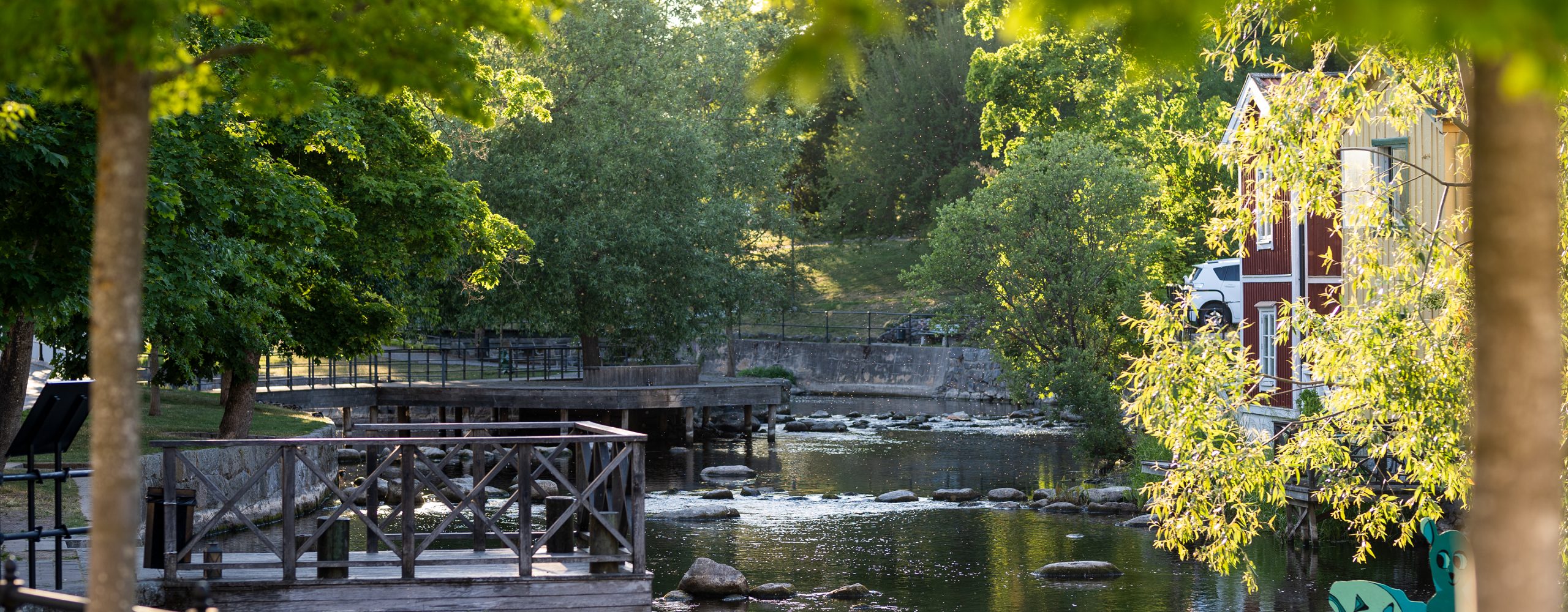 Å med träd, brygga och hus i kvällssol.