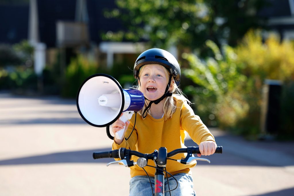 Barn med hjälm cyklar och håller i en megafon utomhus.