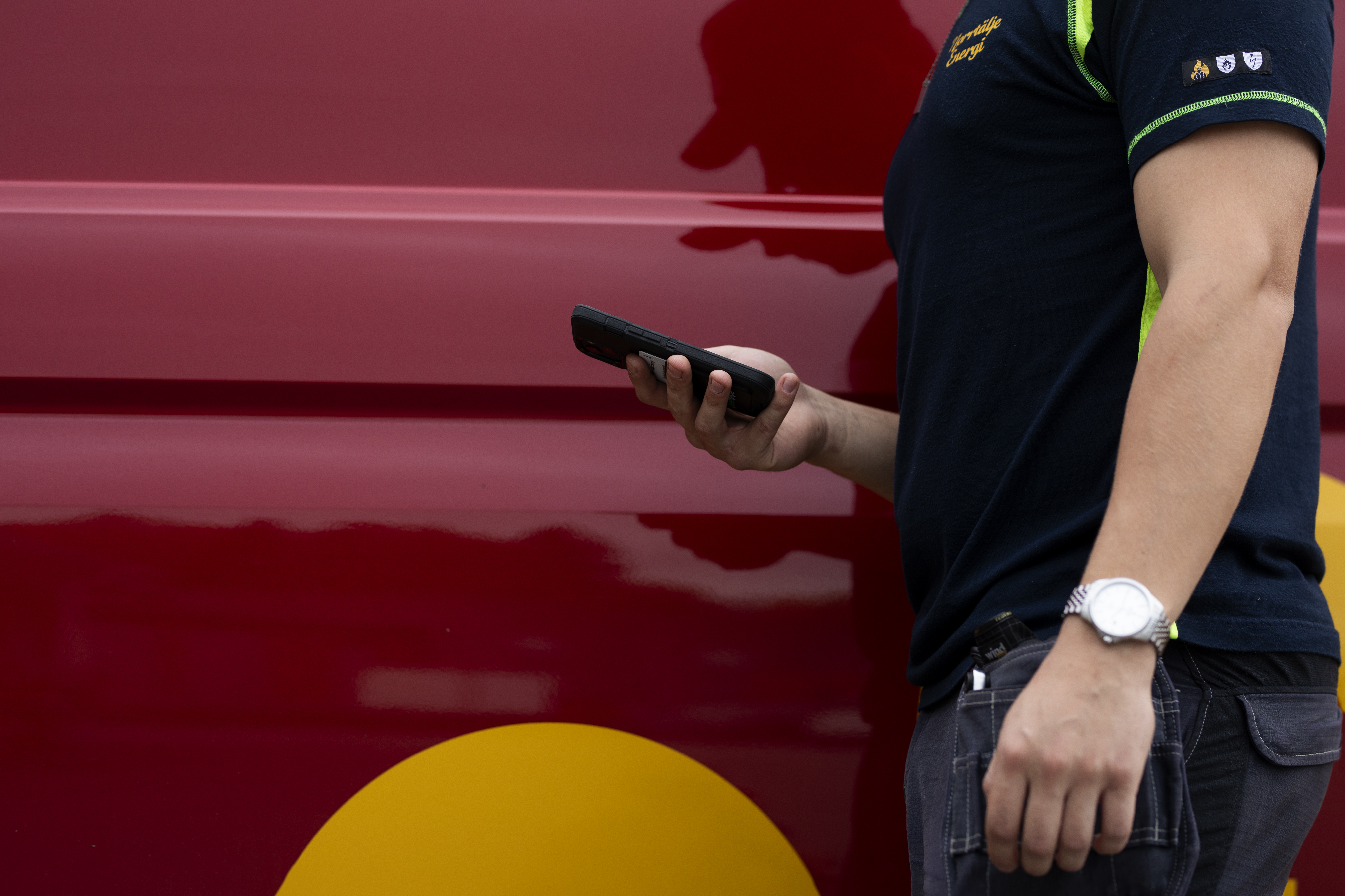 Person i arbetskläder håller en mobiltelefon framför ett rött fordon.