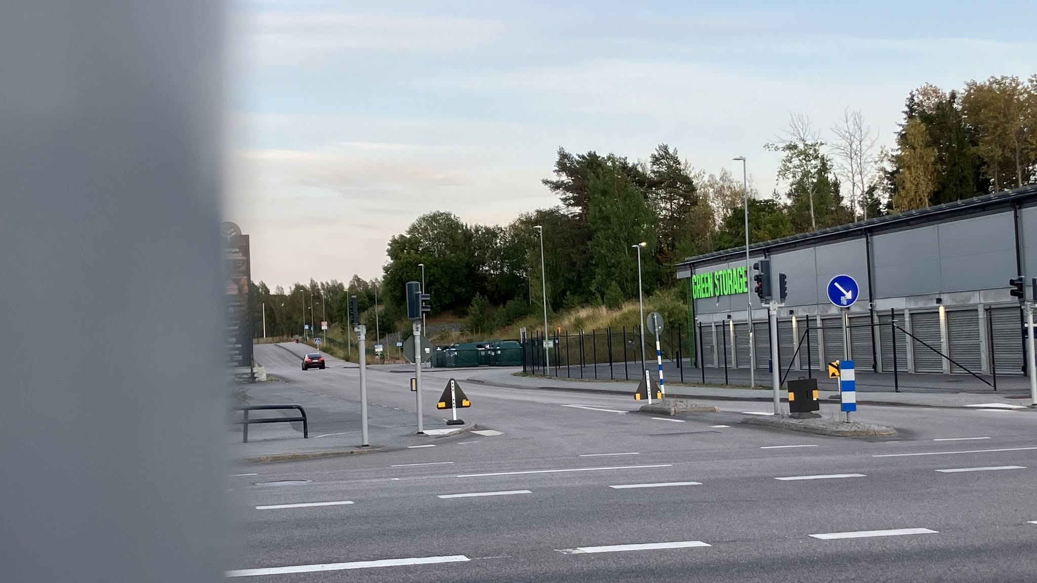 Gatukorsning vid en industribyggnad, med vägmärken, staket och träd i bakgrunden.